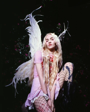 Young woman in fairy costume with feathered wings poses in an outdoor garden with dark lighting and pink flowers in the background.