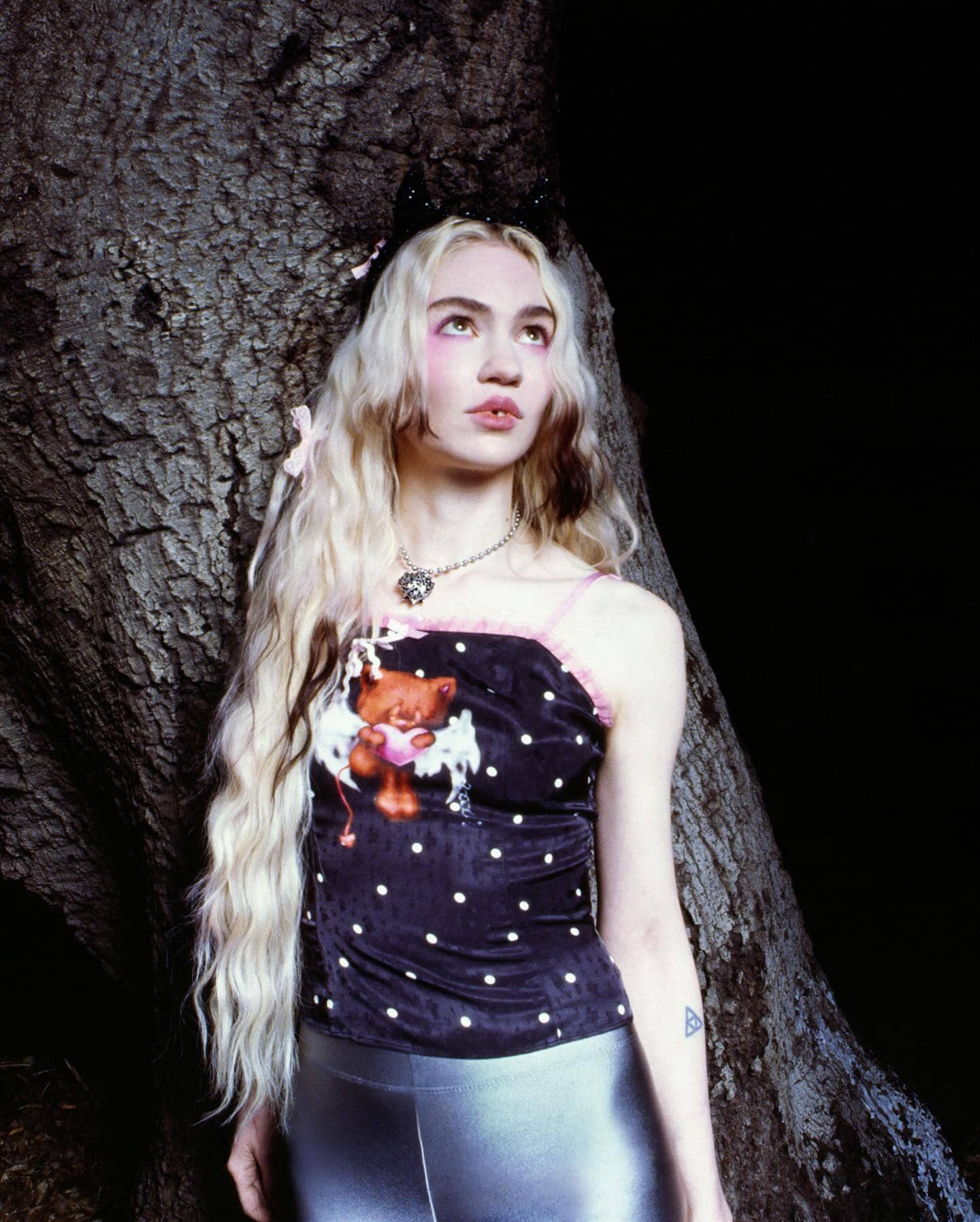 Young adult woman with pale skin and blonde hair poses in the shadows near a tree, wearing a black polka-dot top and silver leggings.