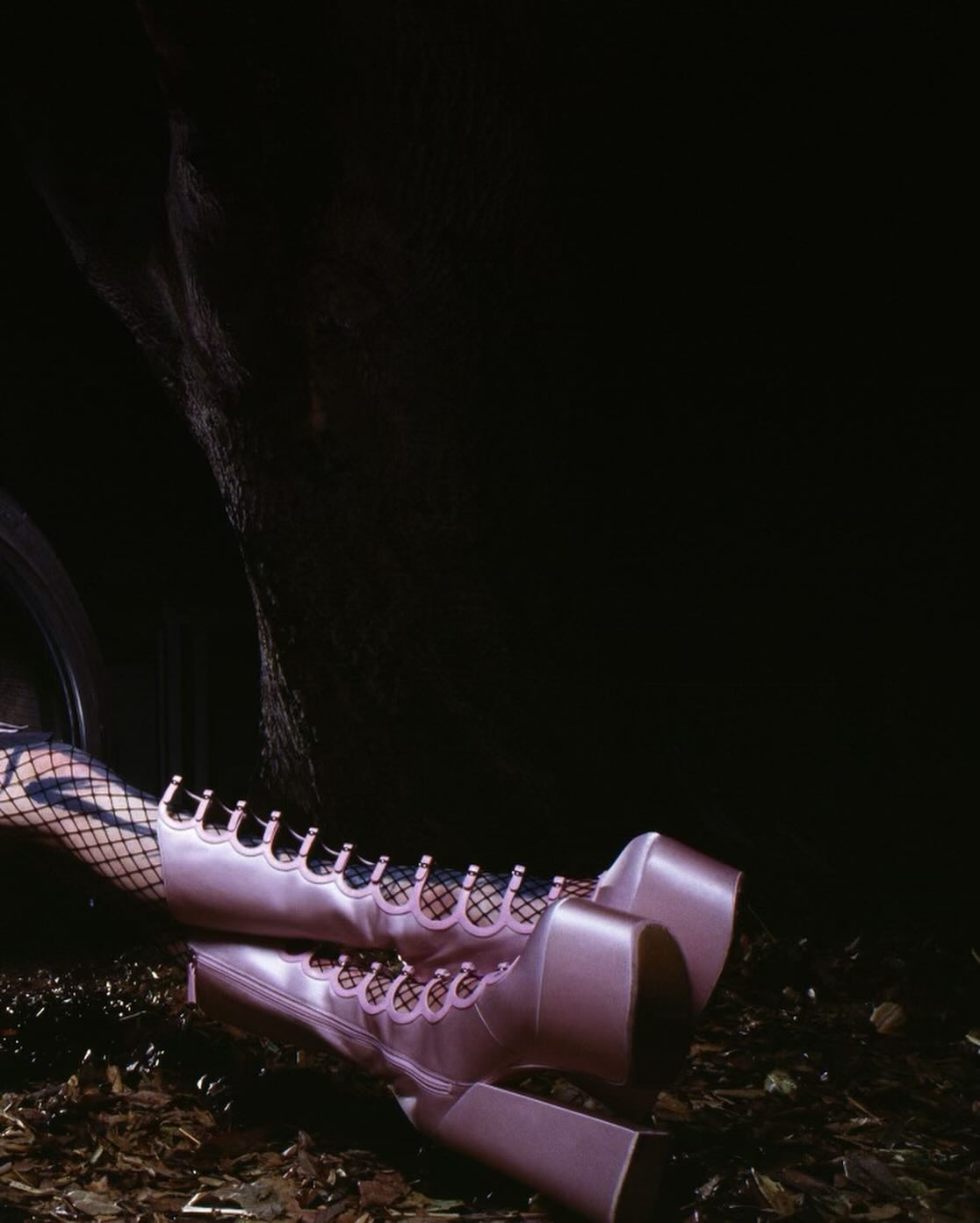 Elevated view of a human wearing light pink platform boots and black fishnet stockings, sitting amidst a dark environment with scattered debris.