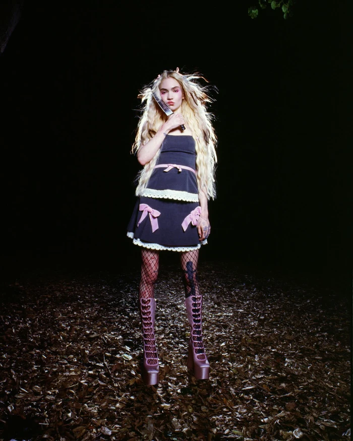 Grimes holding a knife wearing a black dress and platform boots in a dark forest setting for Heaven by Marc Jacobs.