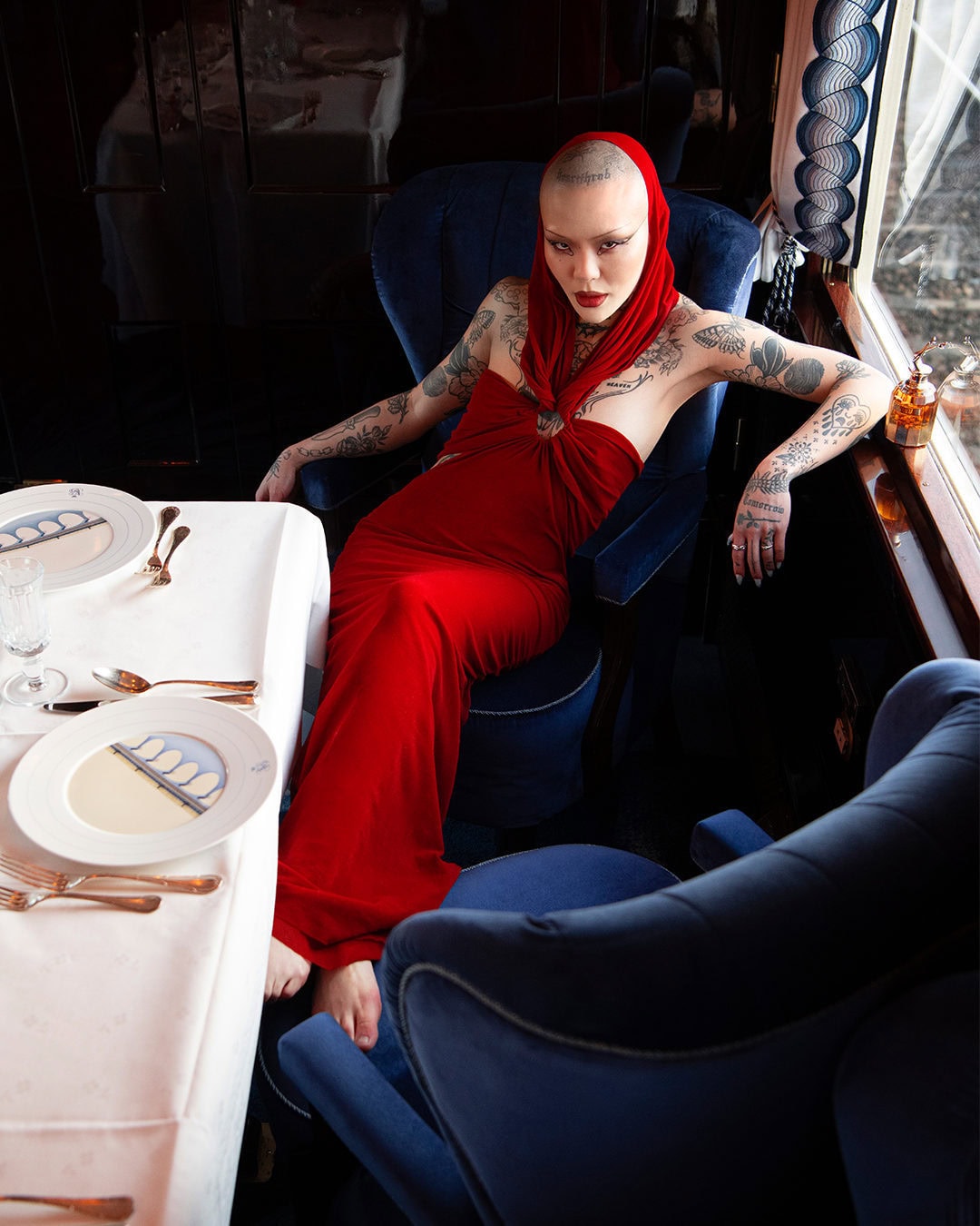 Woman in a red dress seated in a blue chair at a formal dining table set inside a train car, offering a posed portrait.
