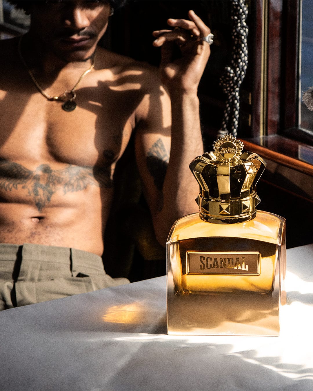 Close-up of a gold Scandal perfume bottle in front of a dark-haired, shirtless man wearing a hat and gold chain necklace.