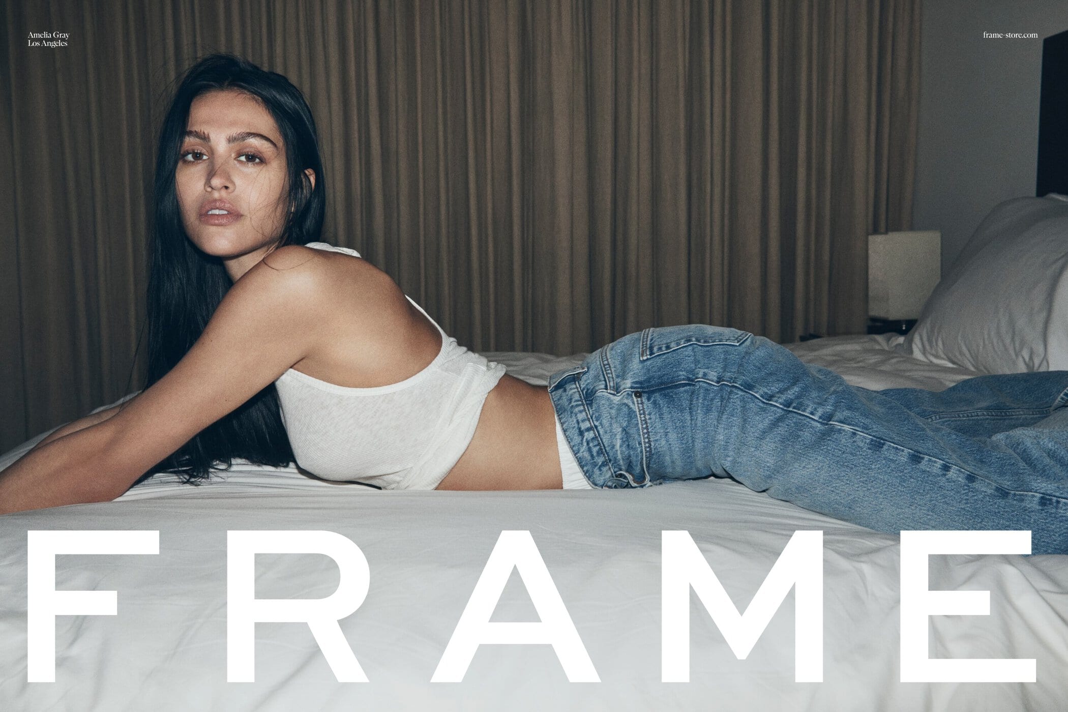 Woman lying on bed wearing white tank top and jeans in a fashion editorial image with soft lighting in an indoor bedroom setting.