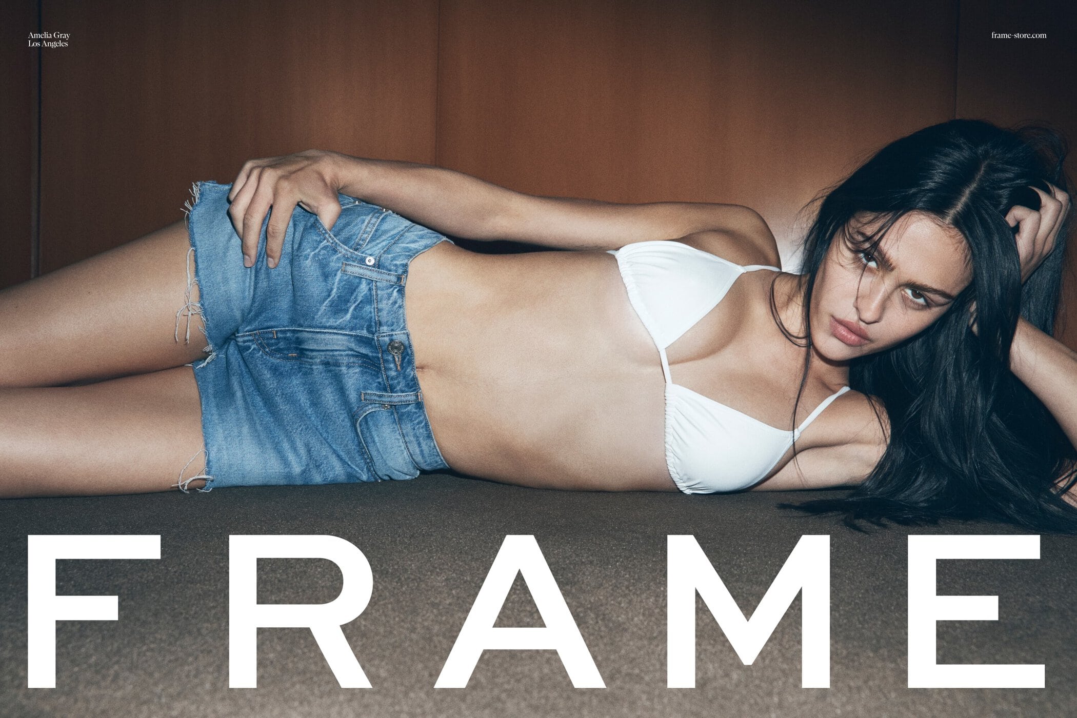 A model lies on a surface wearing a white bikini top and jean shorts with the word 'FRAME' in big white block letters.