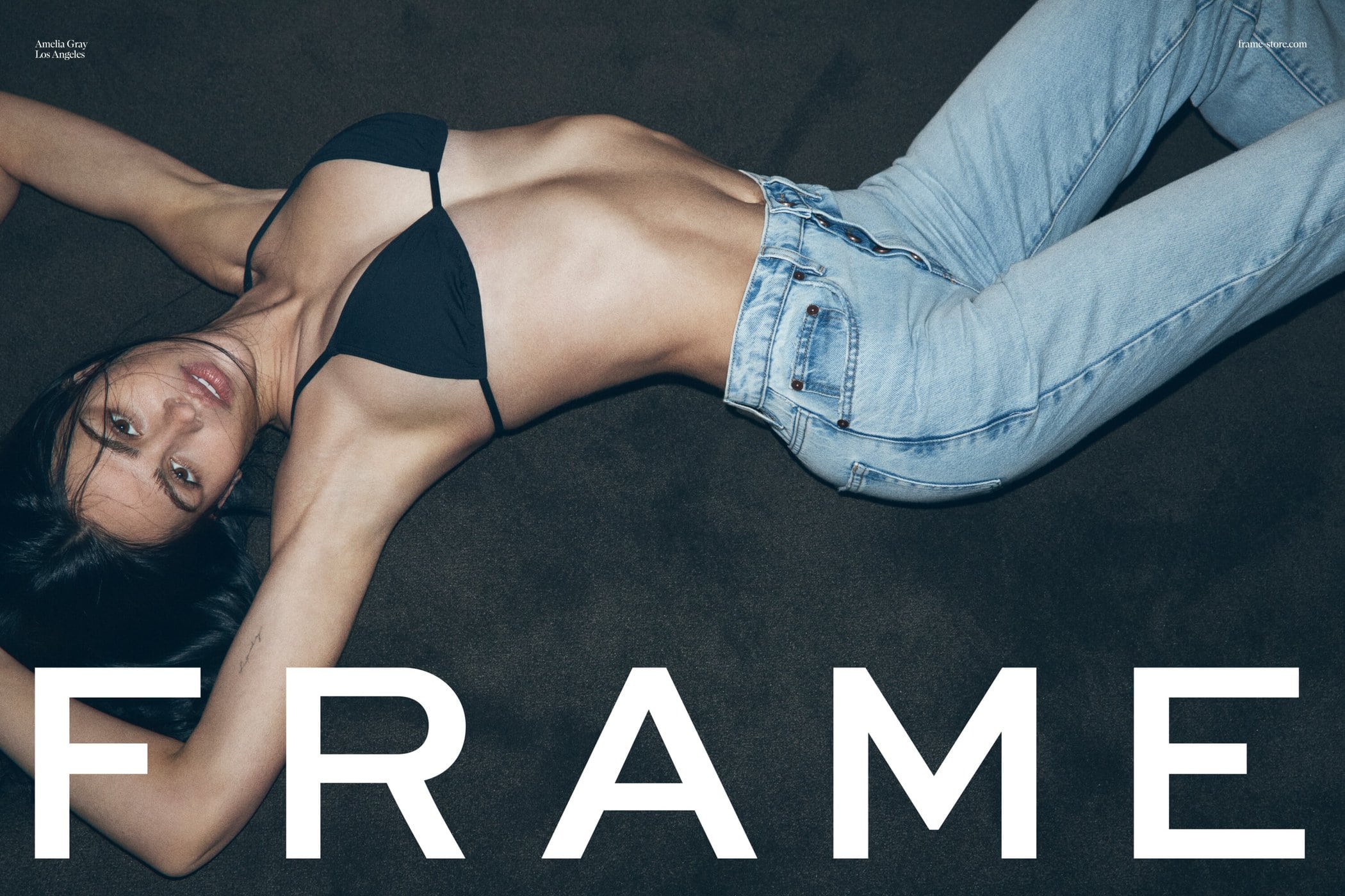 Amelia Gray wearing a black bikini top and light blue denim jeans lying on a dark carpet for FRAME spring 2024 campaign