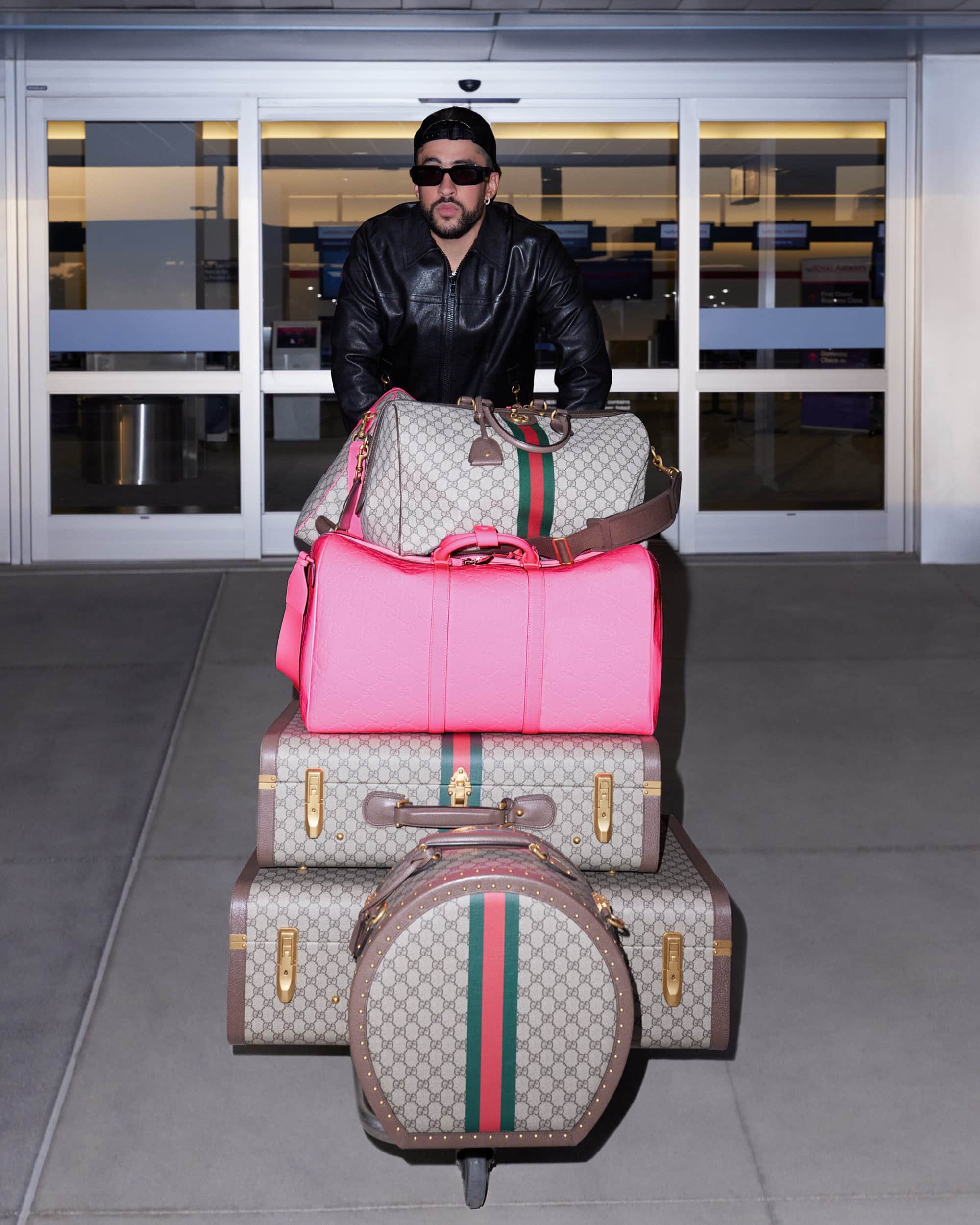 Bad Bunny stands behind a stack of luggage on a luggage cart in front of an airport entrance wearing a black leather jacket and sunglasses.