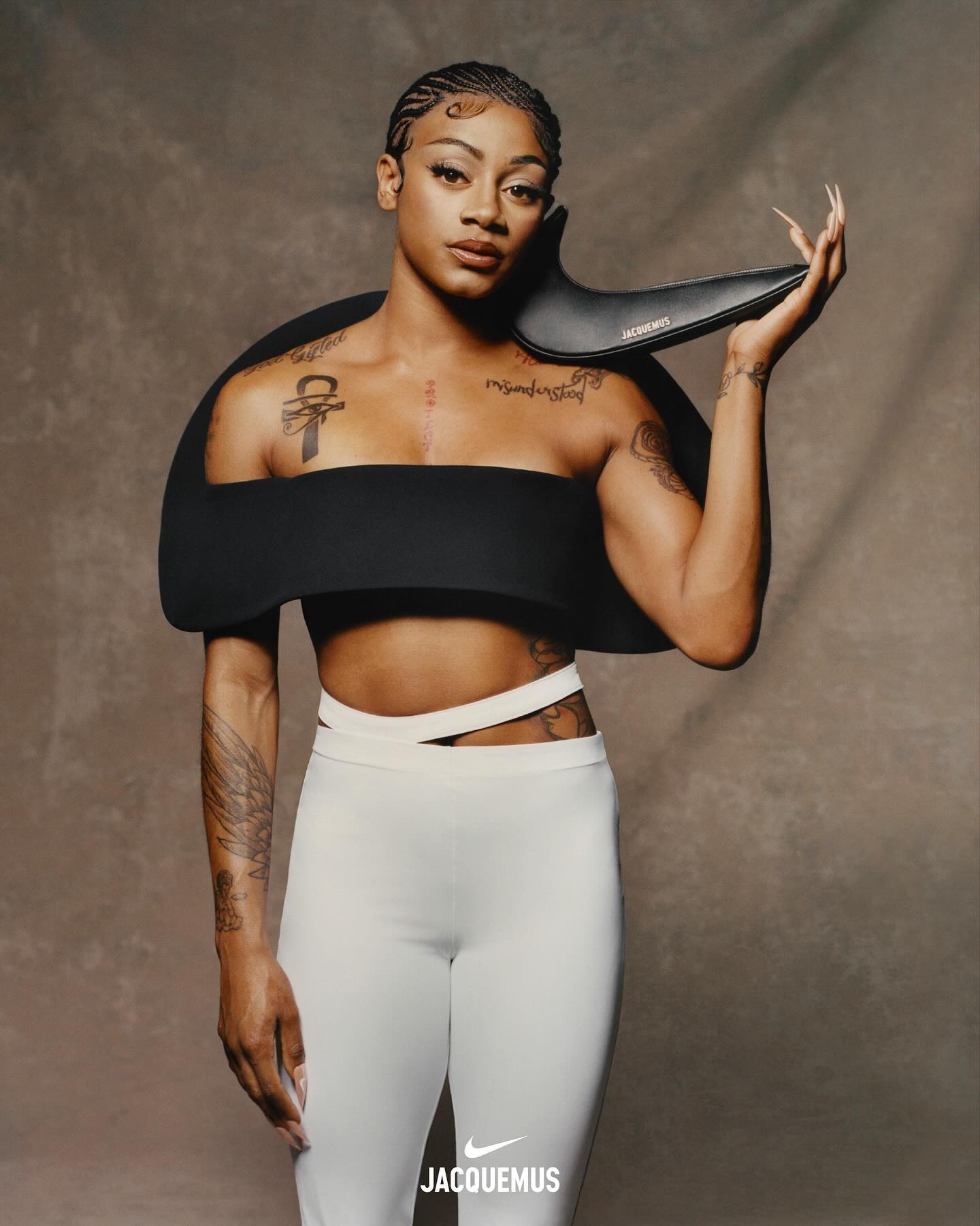 A model poses in a Jacquemus outfit: a black top with open shoulder design, white pants with side cutouts, and logo-branded black accessory.