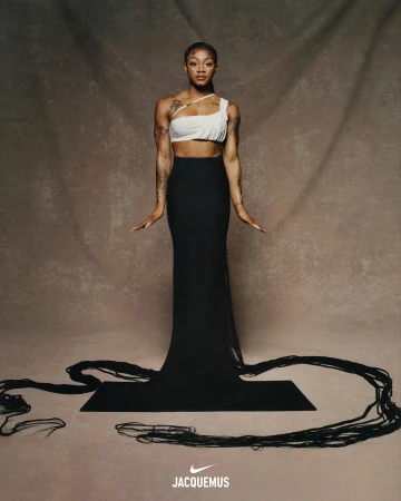 Fashionable woman in white top and black skirt poses on a black yoga mat against a mottled brown background in a studio setting.