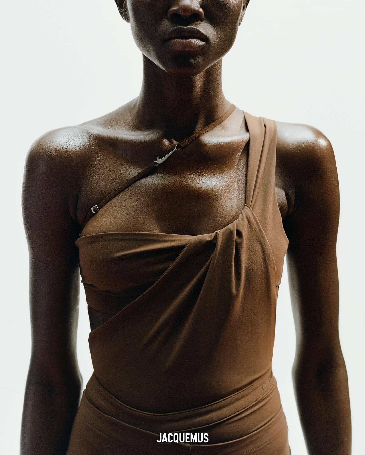 Close-up studio shot of a slender, tan-skinned woman in a draped brown dress looking at the camera against a neutral background.