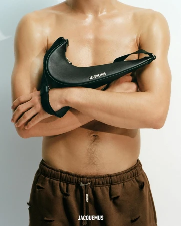 Torso shot of a man holding a black Jacquemus bag. The man has brown torn shorts and is against a neutral background.