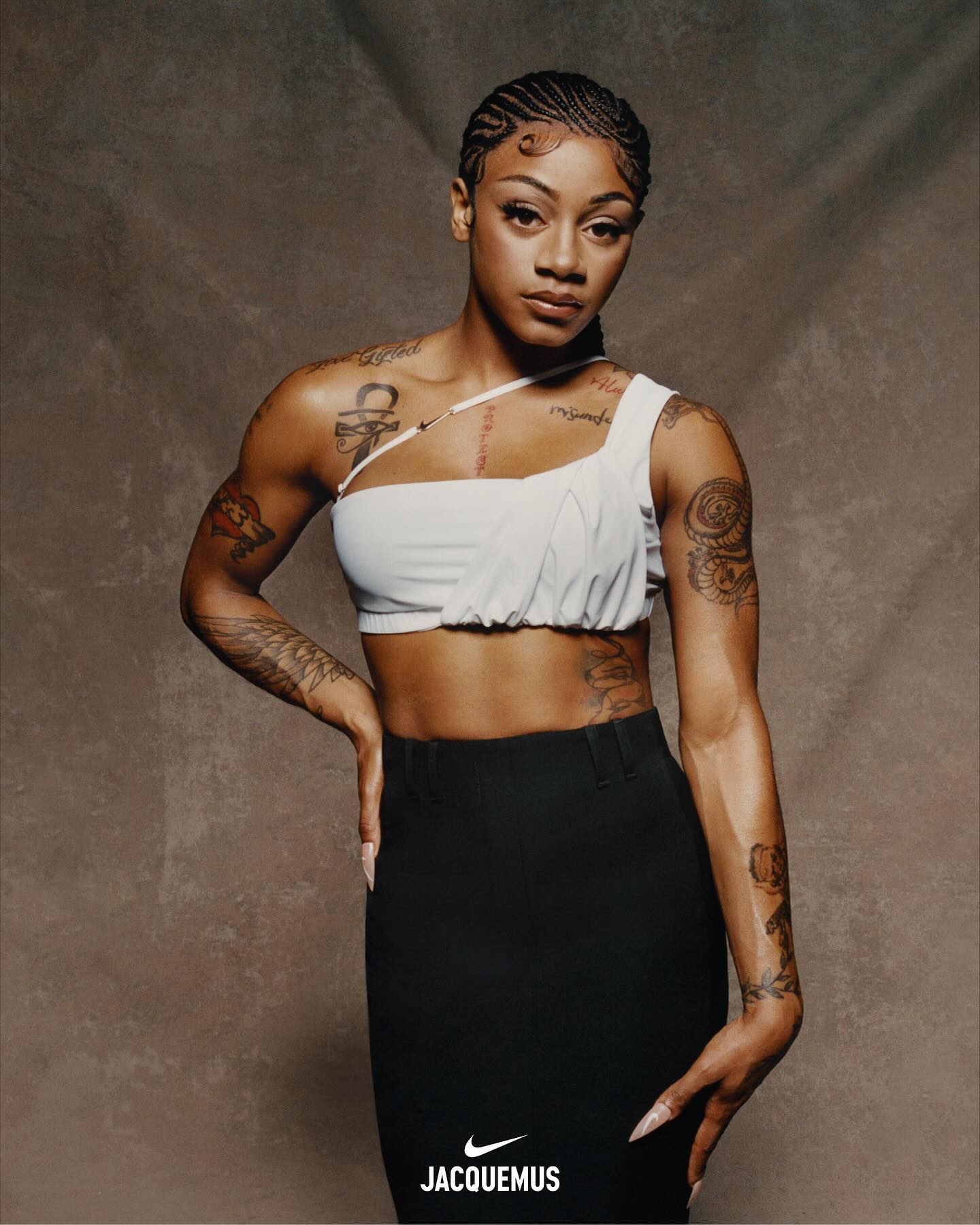 Athletic woman in white crop top and black skirt poses against textured backdrop. Braided hair and tattoos are visible.