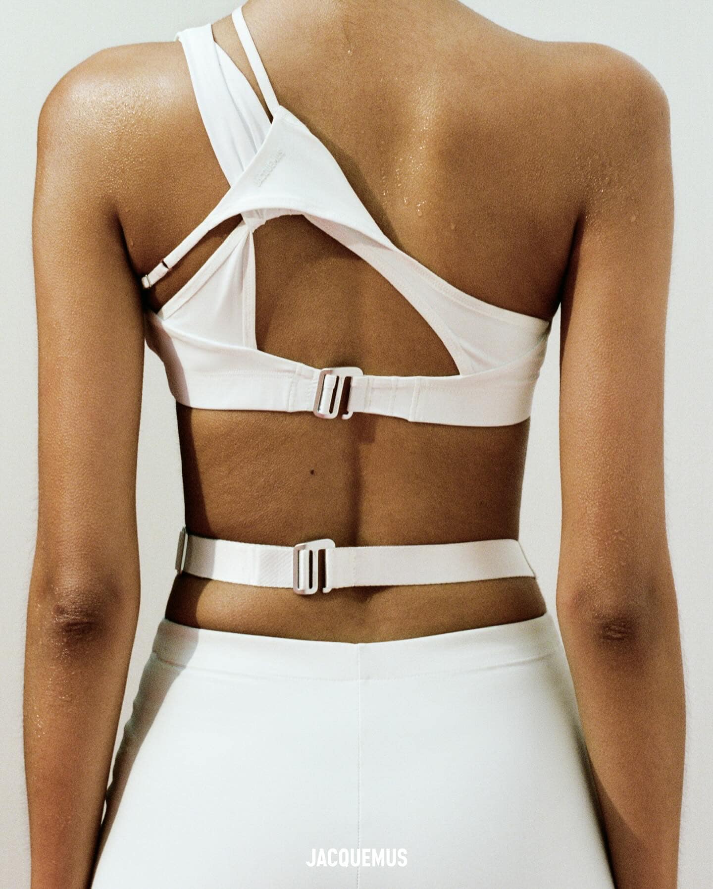 Back view of a tanned-skin adult female wearing a white sports bra with cut-out detailing and matching athletic pants. Indoor setting.