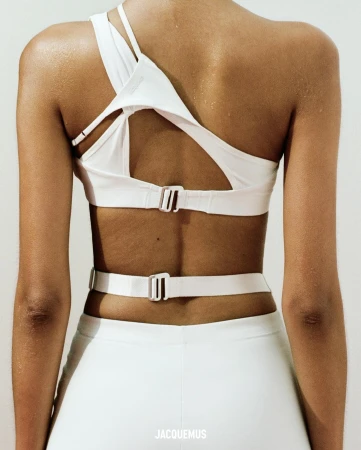 Back view of a tanned-skin adult female wearing a white sports bra with cut-out detailing and matching athletic pants. Indoor setting.