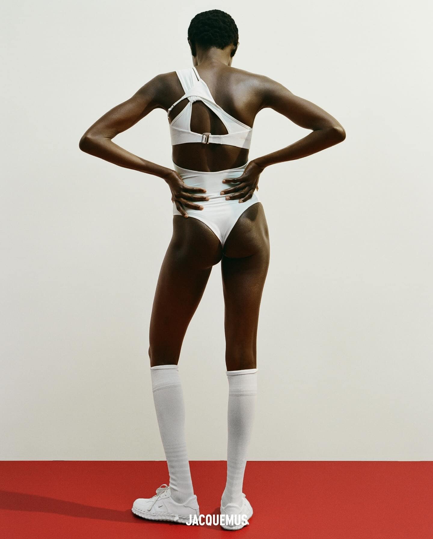 Back view of a Black female fashion model in a white one-shoulder bathing suit, white socks and white shoes, standing on a red surface.