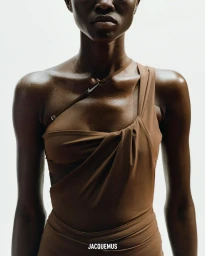 Close-up of a model wearing a brown asymmetric one-shoulder top against a plain white background for Jacquemus and Nike.