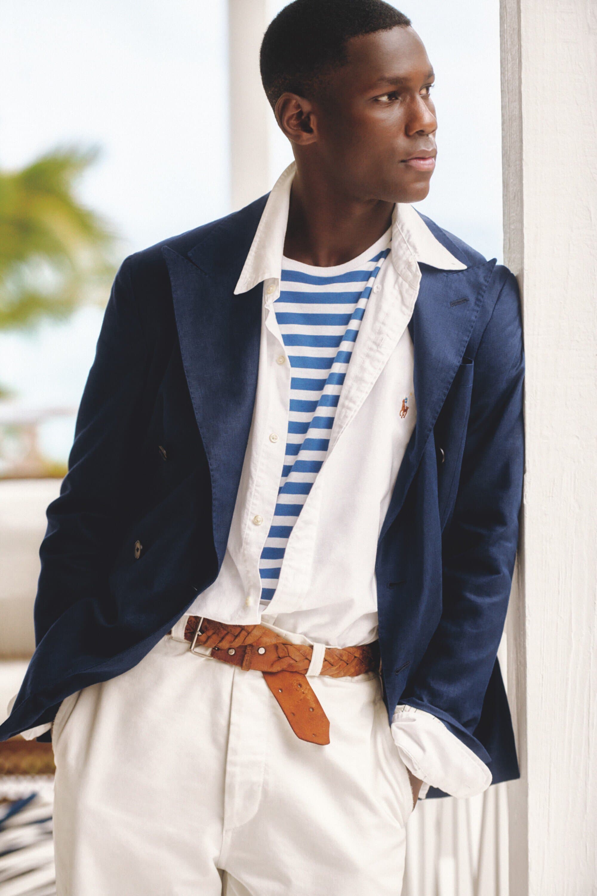 Adult man in a navy blazer poses in front of a white wall. His shirt has blue stripes and he's wearing white pants and a brown belt.