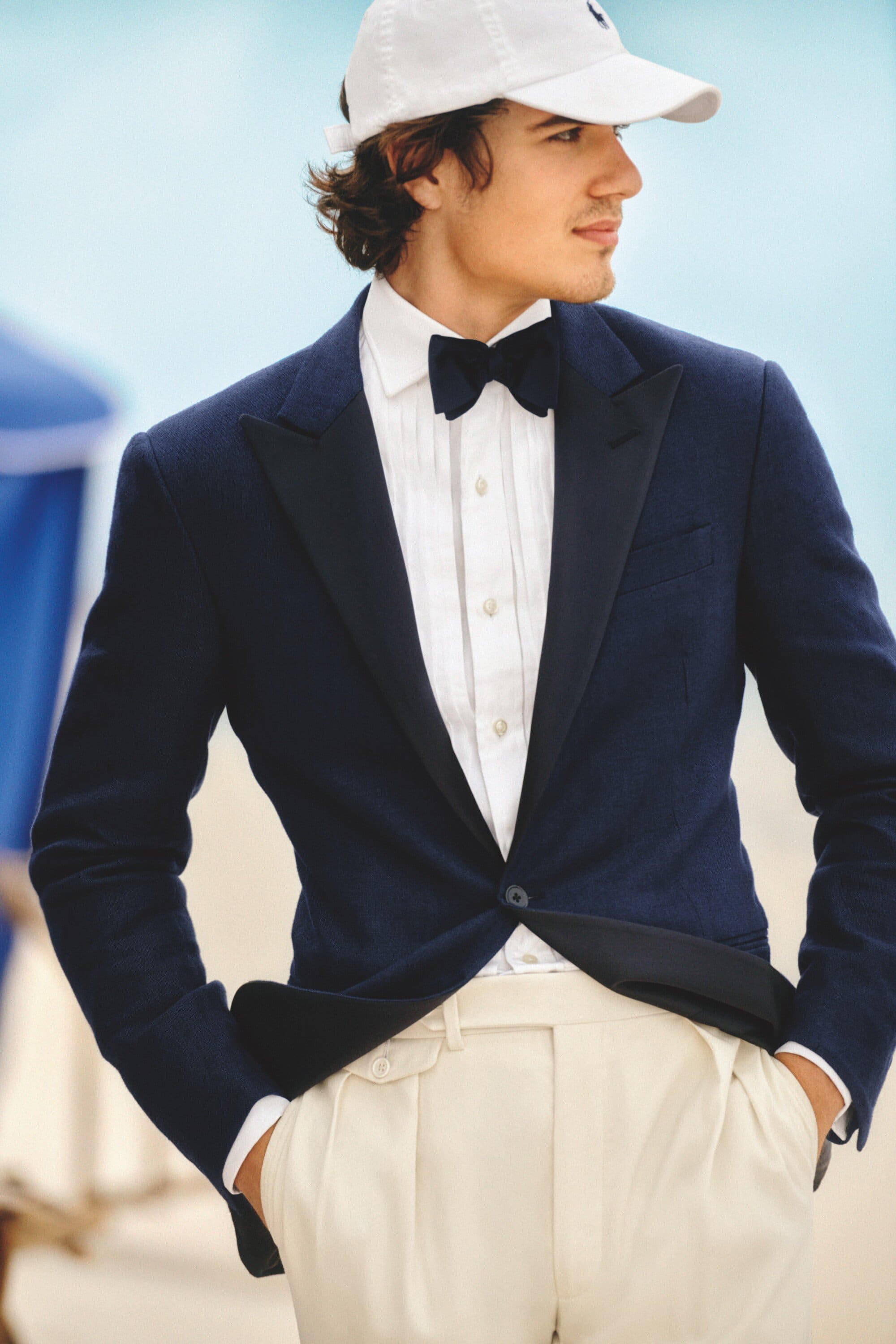 Man in navy tuxedo jacket, white shirt, bow tie, and baseball cap, outdoors with soft blue background, looking to the side.