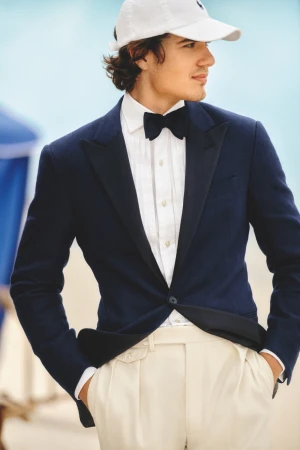 Man in navy tuxedo jacket, white shirt, bow tie, and baseball cap, outdoors with soft blue background, looking to the side.