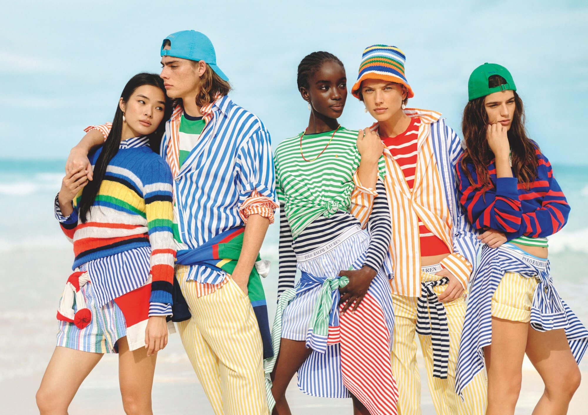 Five fashion models in colorful striped tops and striped shorts or pants pose on a sandy beach with a light blue sky and ocean backdrop.