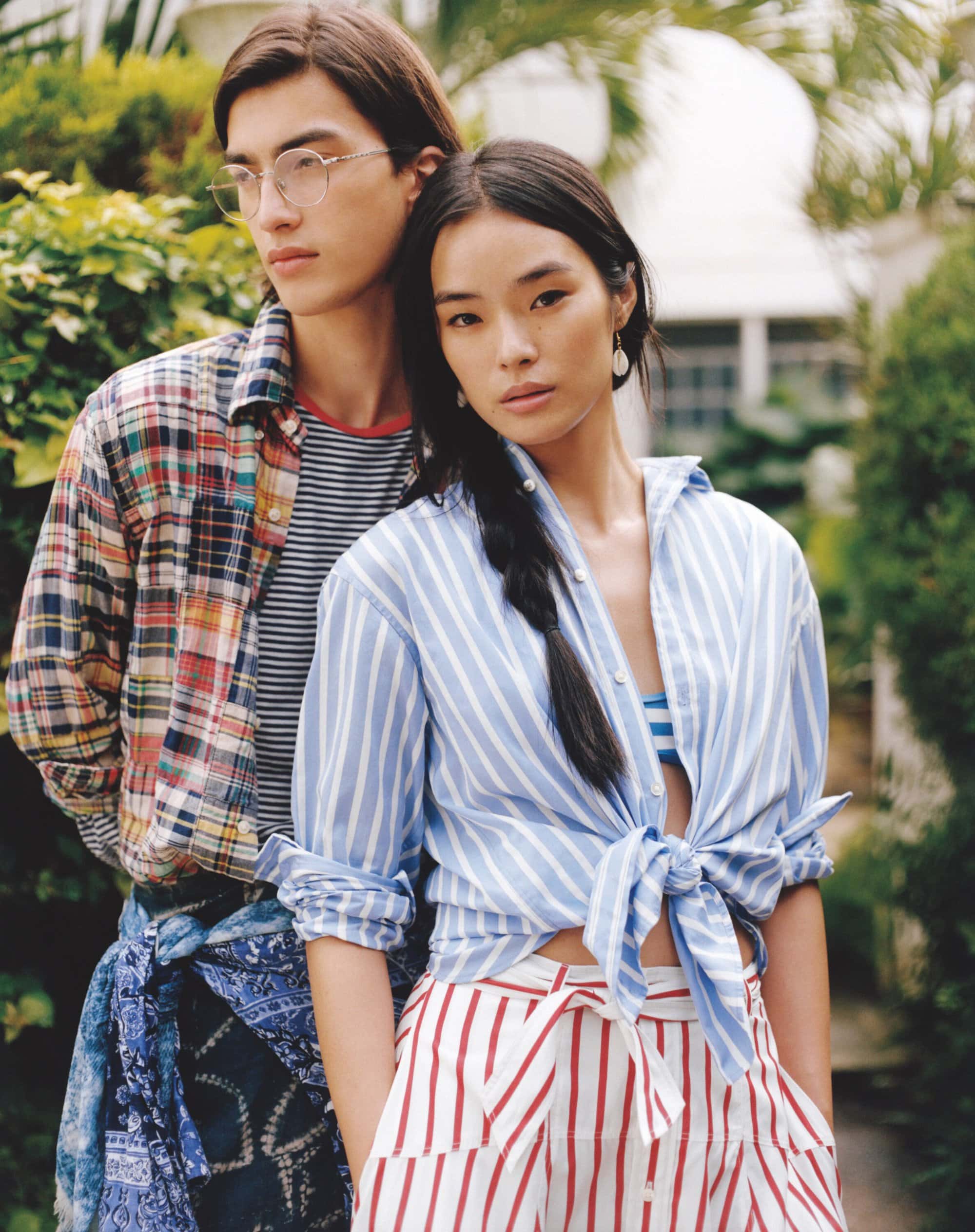 Two models, an adult male and an adult female, pose in an outdoor setting wearing casual striped shirts, shorts, and a plaid shirt.