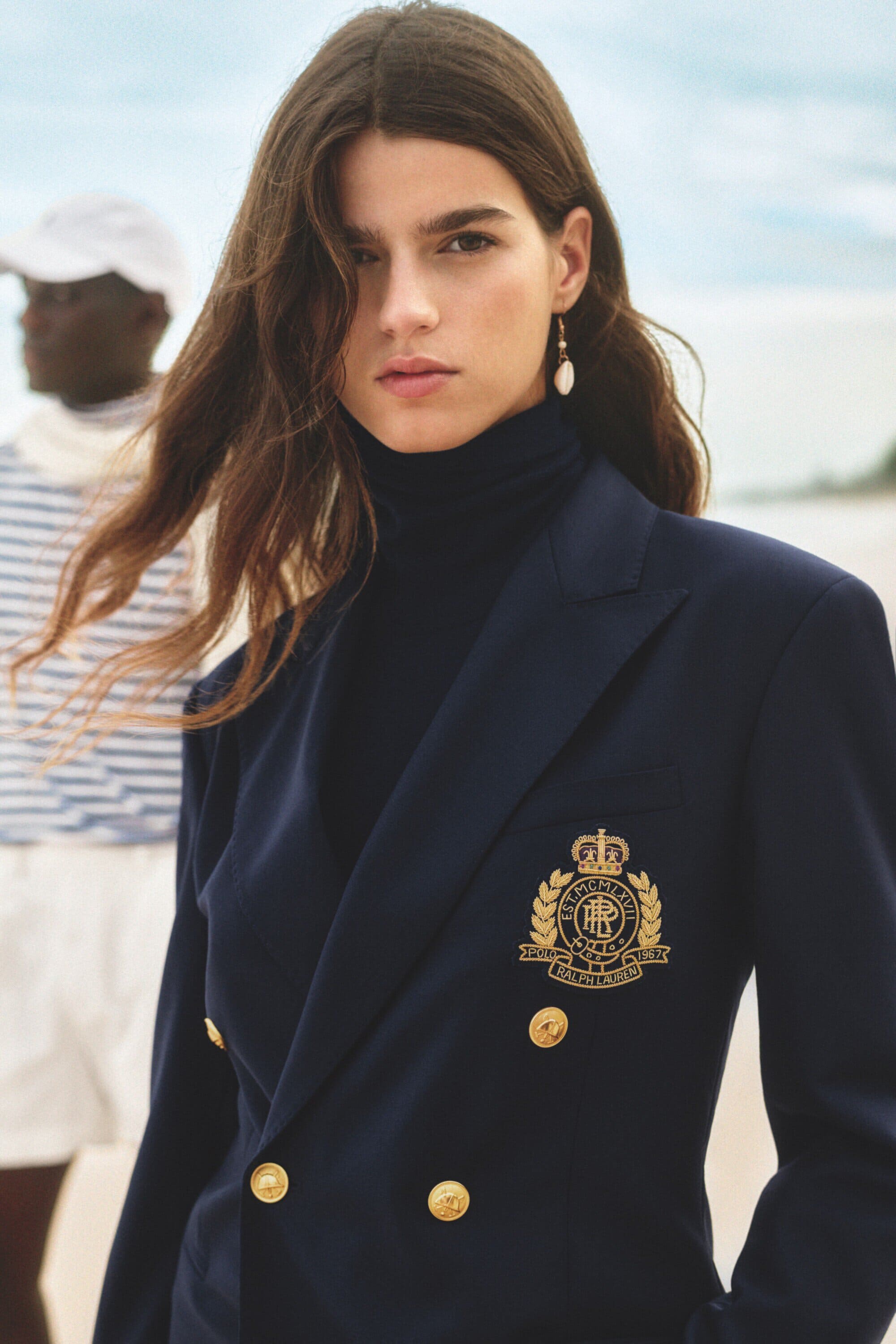 Fashion shot of a woman in a navy blazer with gold buttons and a black turtleneck, standing on a beach with another person behind.