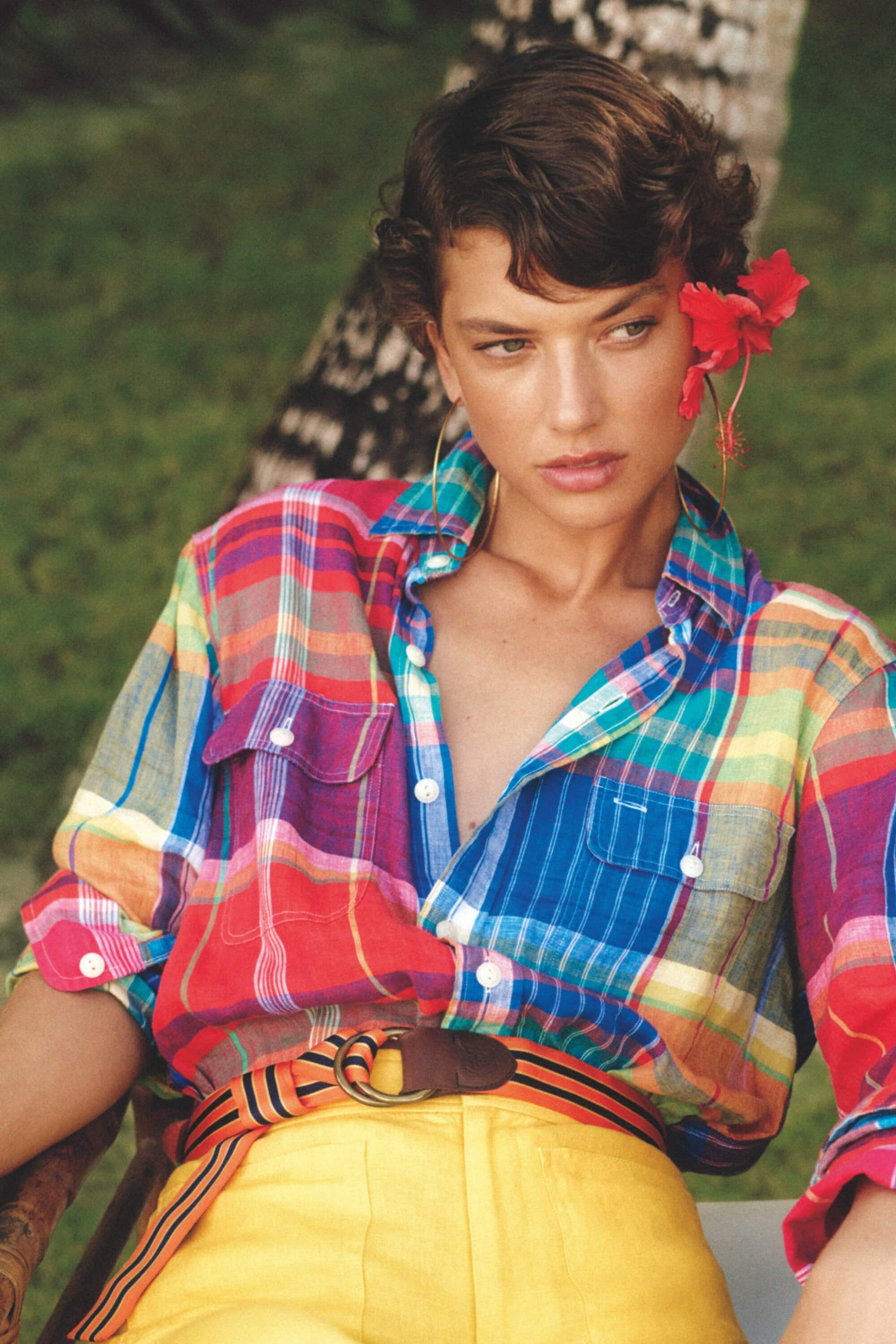 Portrait of a brunette woman in a colorful plaid shirt and yellow shorts with a striped belt, accessorized with a red flower and gold hoops.