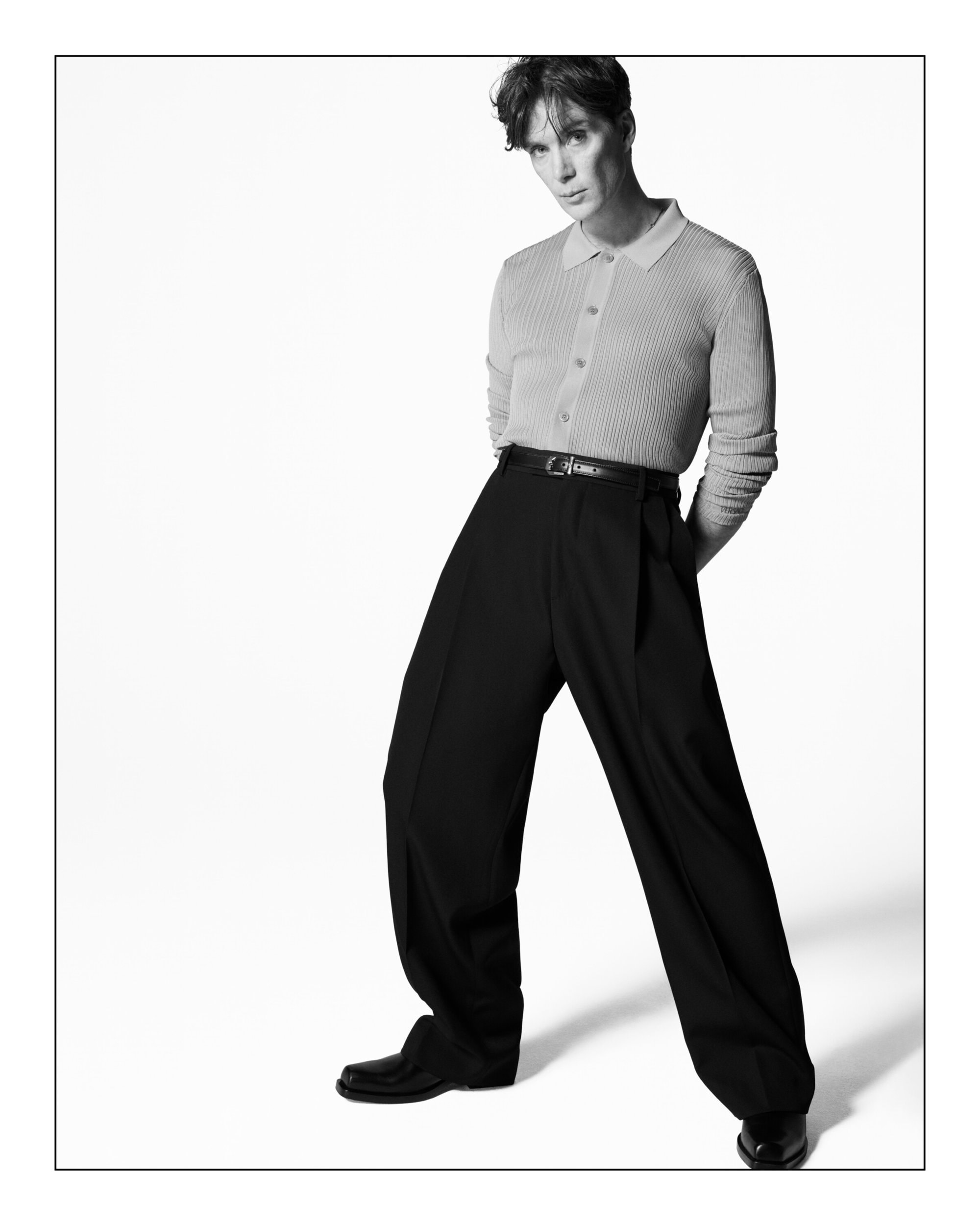 Monochrome full shot of an adult man with a neutral expression, wearing a light-colored ribbed shirt and dark, wide-legged pants in a studio.