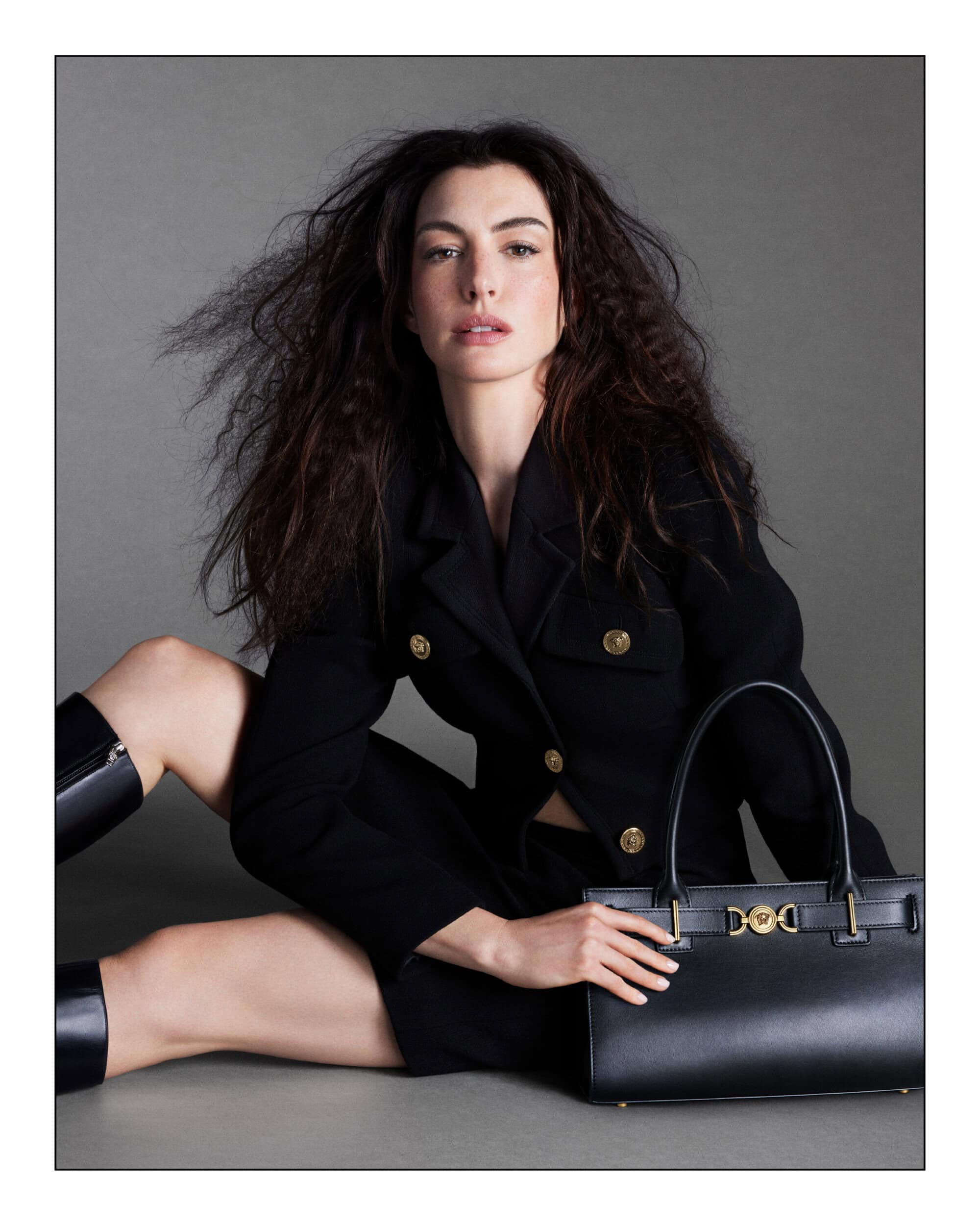 Adult woman in black blazer and skirt sitting on the floor, holding a black purse, with dark wavy hair against a grey backdrop.