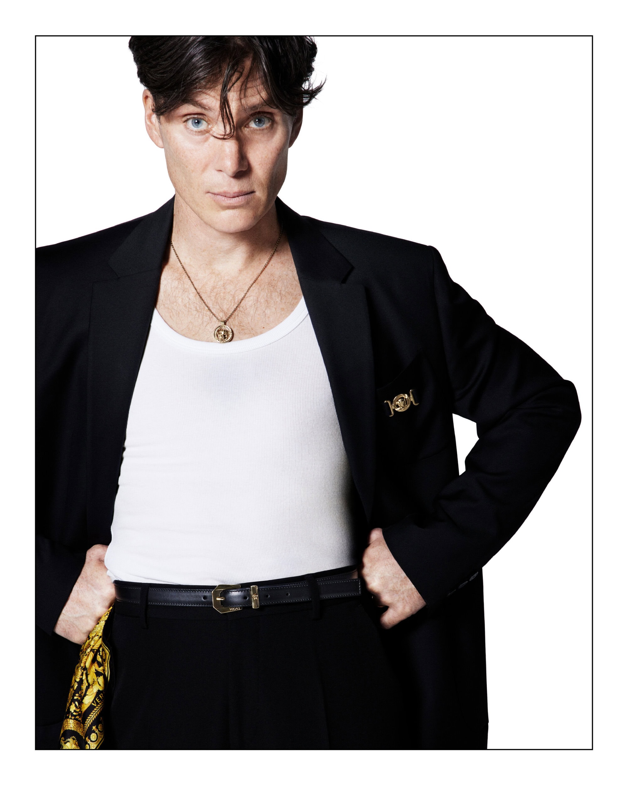 Cillian Murphy in a black suit jacket and white tank top, hands on hips, stares directly at the viewer in a studio setting.