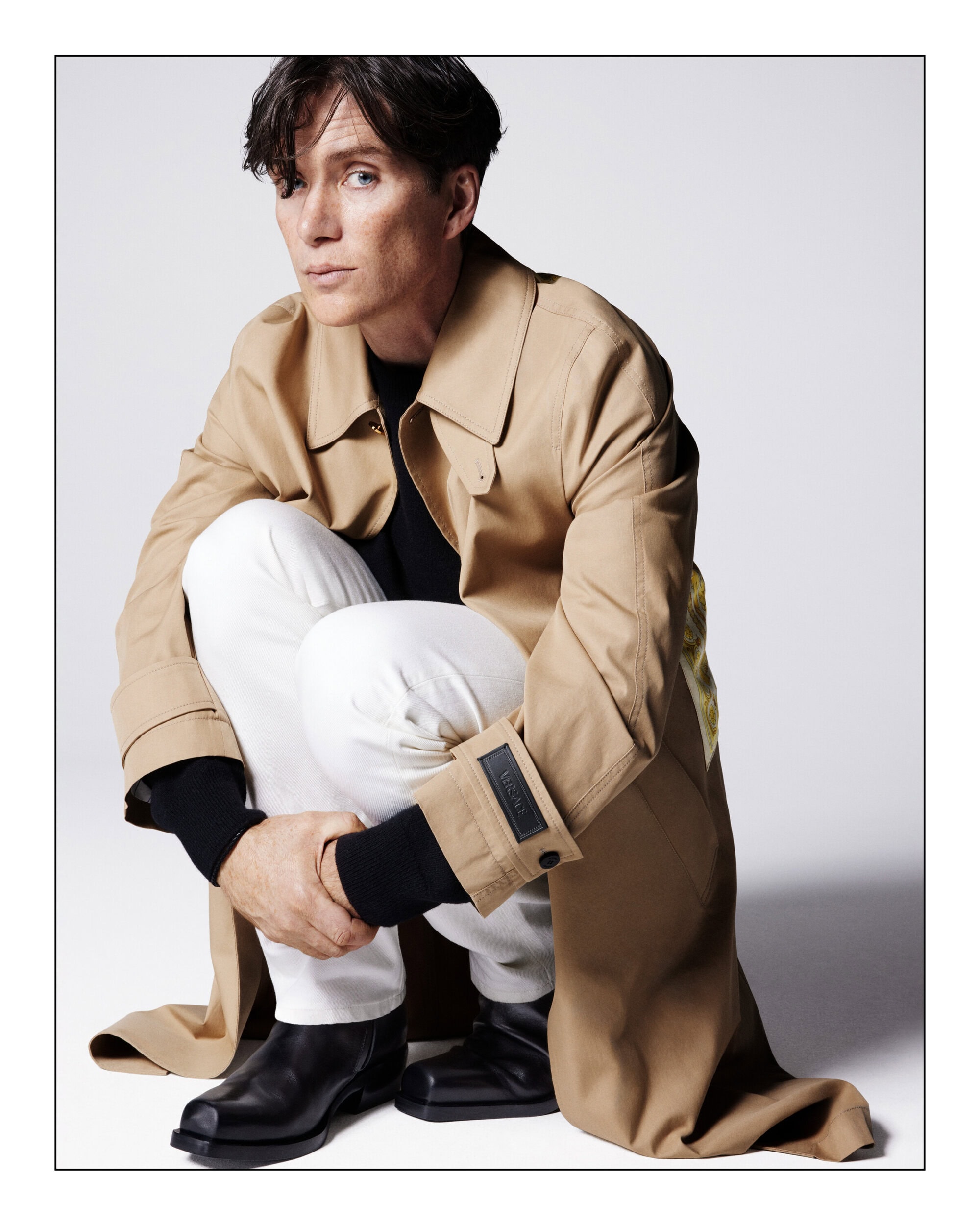 A man in a tan trench coat poses against a white backdrop, seated with knees up, showcasing black top and shoes, and white pants.