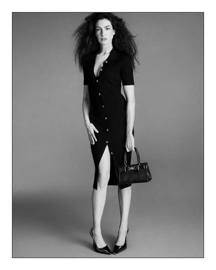 Monochrome fashion campaign image of Anne Hathaway wearing a black ribbed midi dress and holding a top-handle handbag against a grey backdrop