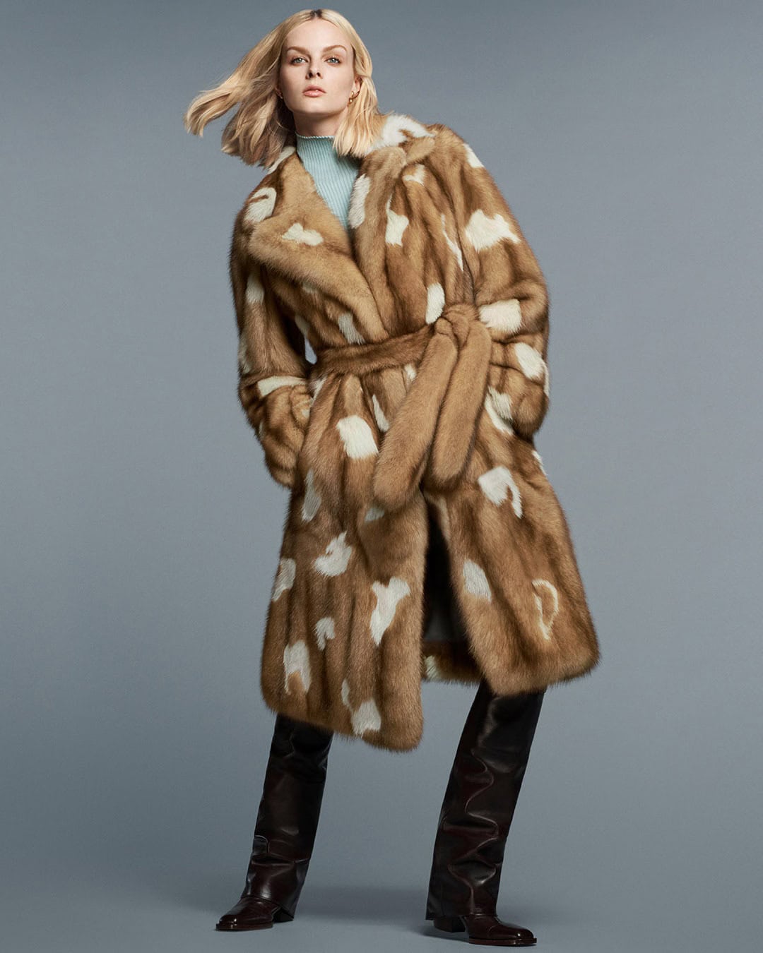 Fashion model poses in studio wearing fur coat, turtle neck sweater, and leather boots against a grey background.