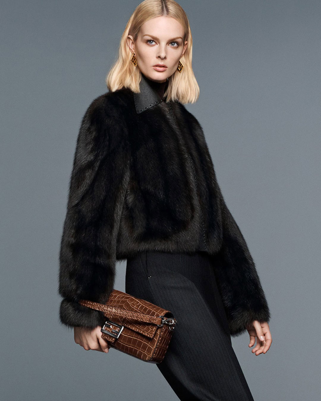 Fashion model wearing a fur coat and holding a brown purse in a studio setting. She has blonde hair and is wearing gold earrings.