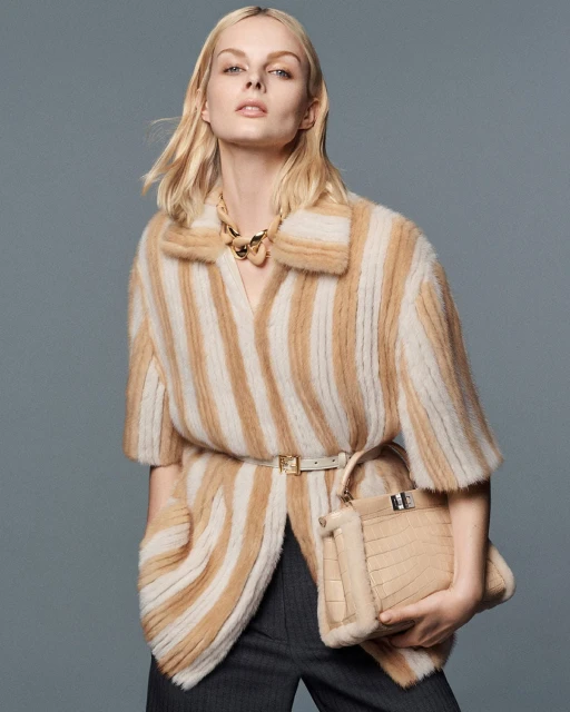 Fashionable woman in a striped coat and gray pants holds a purse in a studio setting, showcasing modern style and elegant design.