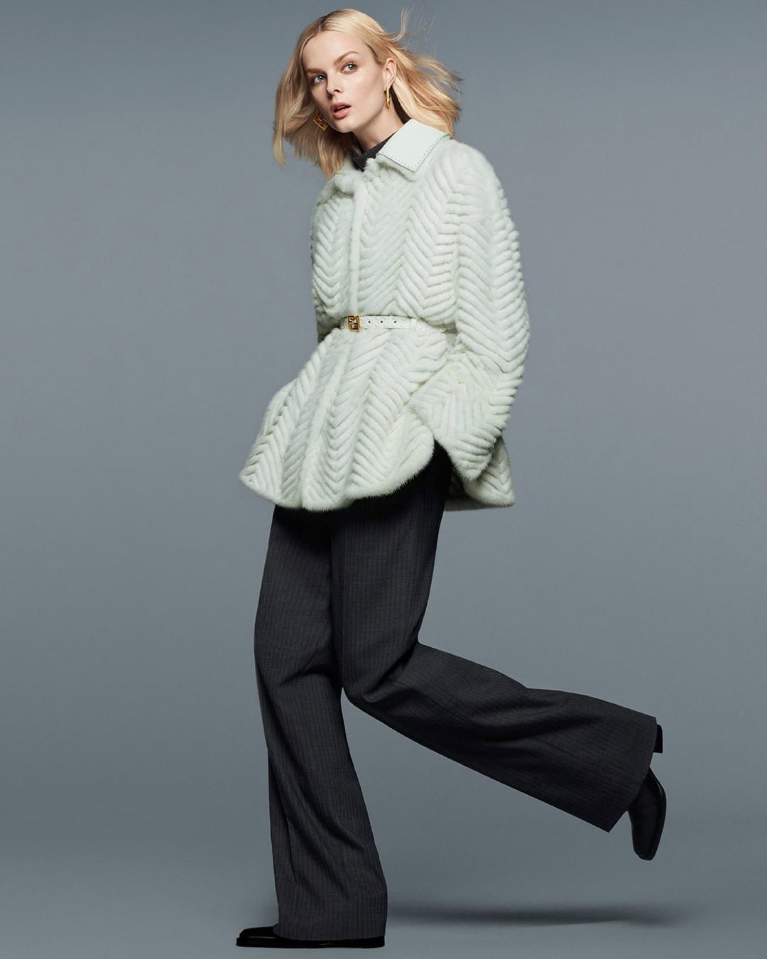 Fashion portrait of a woman in an textured white jacket, grey pinstripe trousers, and black shoes, set against a neutral grey backdrop.
