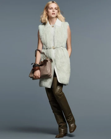 Model Aylah Peterson wearing a pale green fur vest and olive leather boots holding a brown handbag against a grey backdrop