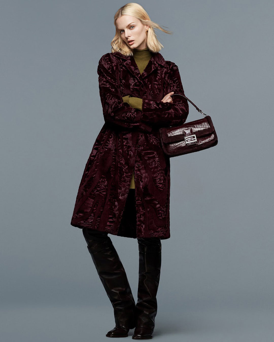 Fashion shot of a woman in a patterned wine-colored coat, dark knee-high boots, and a brown purse in a neutral studio.