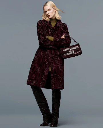 Fashion shot of a woman in a patterned wine-colored coat, dark knee-high boots, and a brown purse in a neutral studio.
