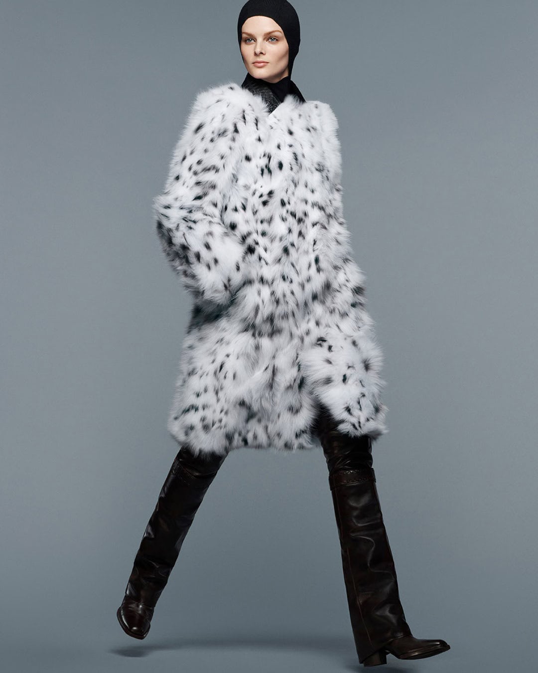 A fashion shot of a model in a black head covering, striking a walking pose in a spotted white fur coat and black, thigh-high leather boots.