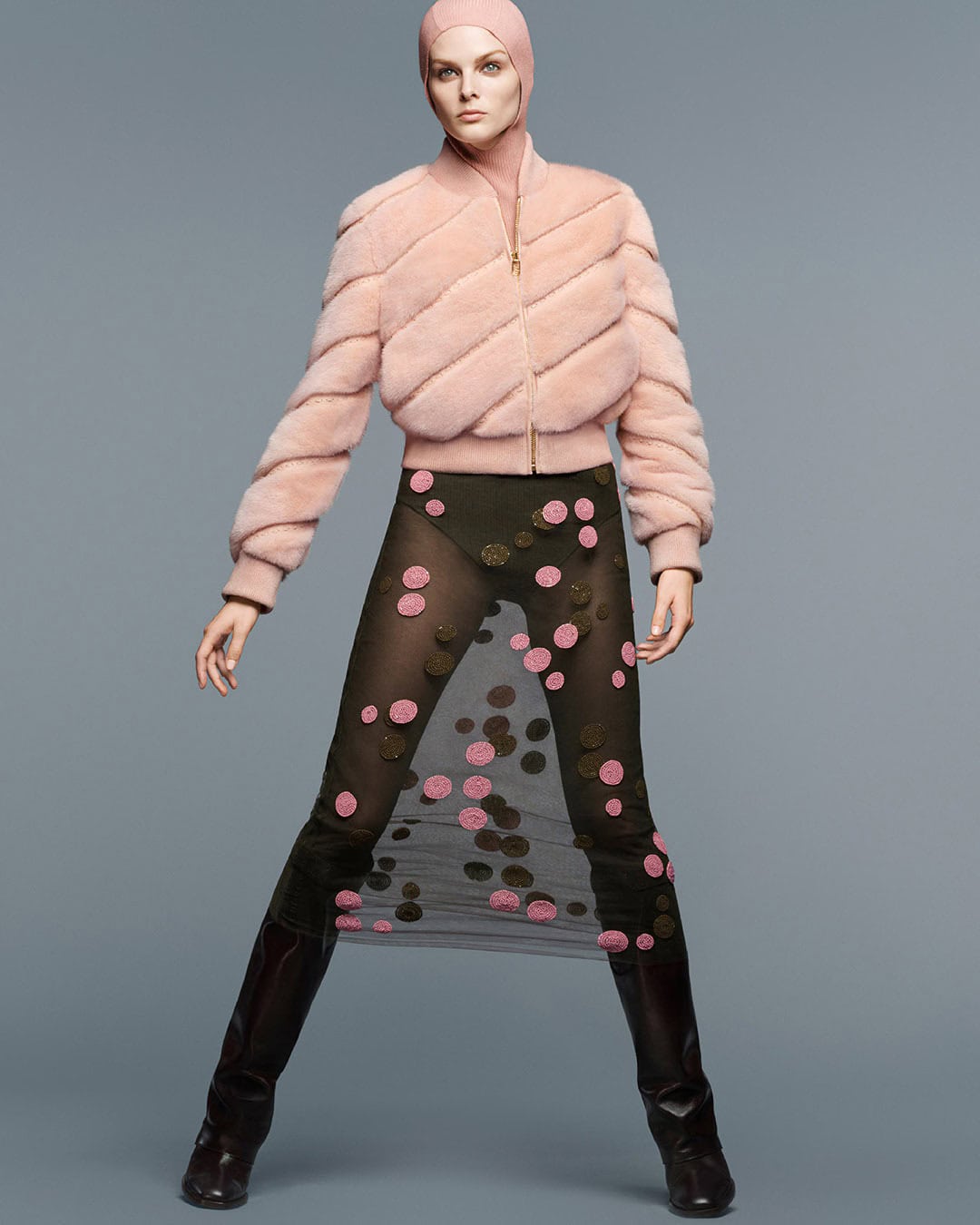 A fashion model poses in a pink jacket, olive green bodysuit and black boots against a neutral backdrop in a full-length shot.