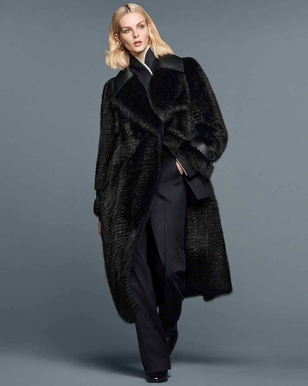 Fashionable woman wearing a long black fur coat and black pants, standing against a grey studio backdrop in a full-length shot.