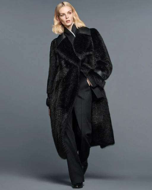 Fashionable woman wearing a long black fur coat and black pants, standing against a grey studio backdrop in a full-length shot.