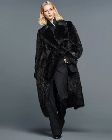 Fashionable woman wearing a long black fur coat and black pants, standing against a grey studio backdrop in a full-length shot.