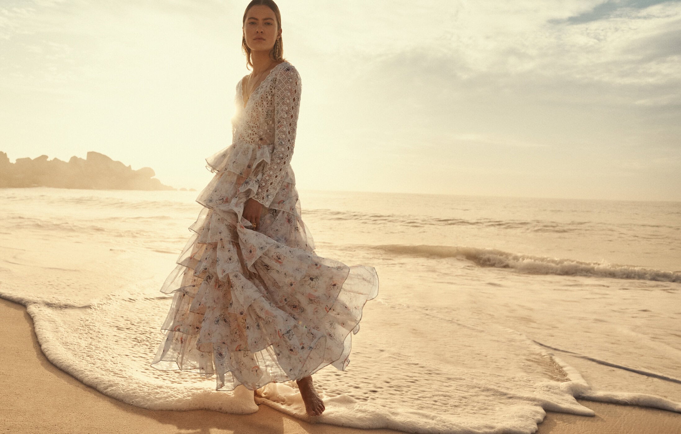 Woman in a flowing floral dress walking along a sandy beach at sunrise, with soft lighting and the ocean in the background.