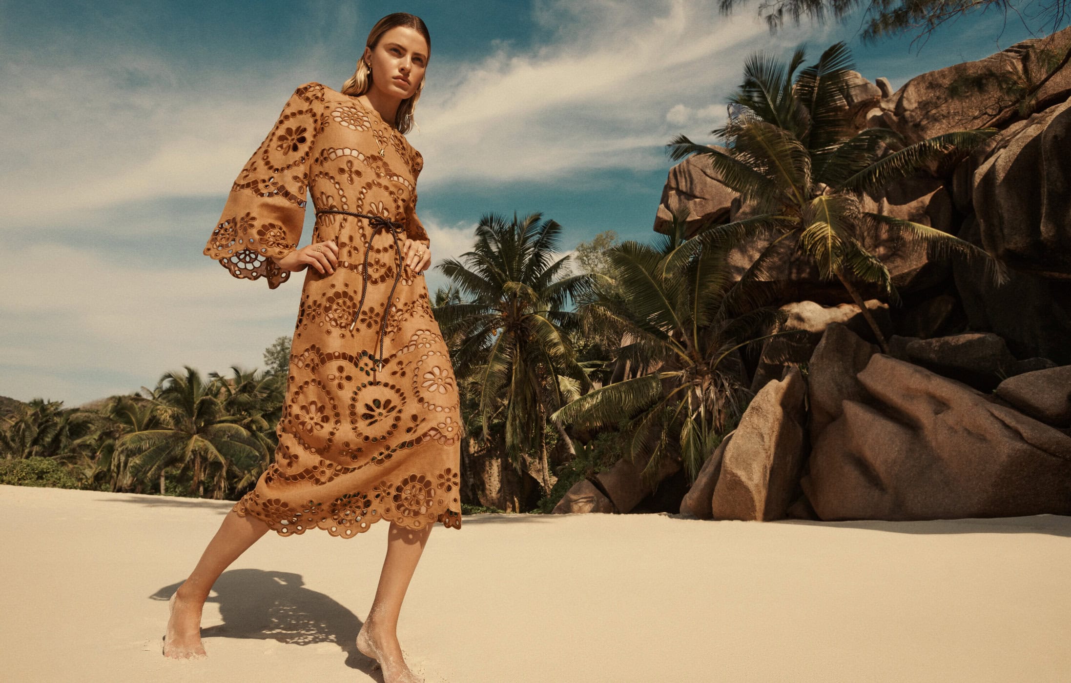 A woman in a tan, patterned dress with a black belt walks barefoot on a sandy beach, backed by tropical trees and rock formations.