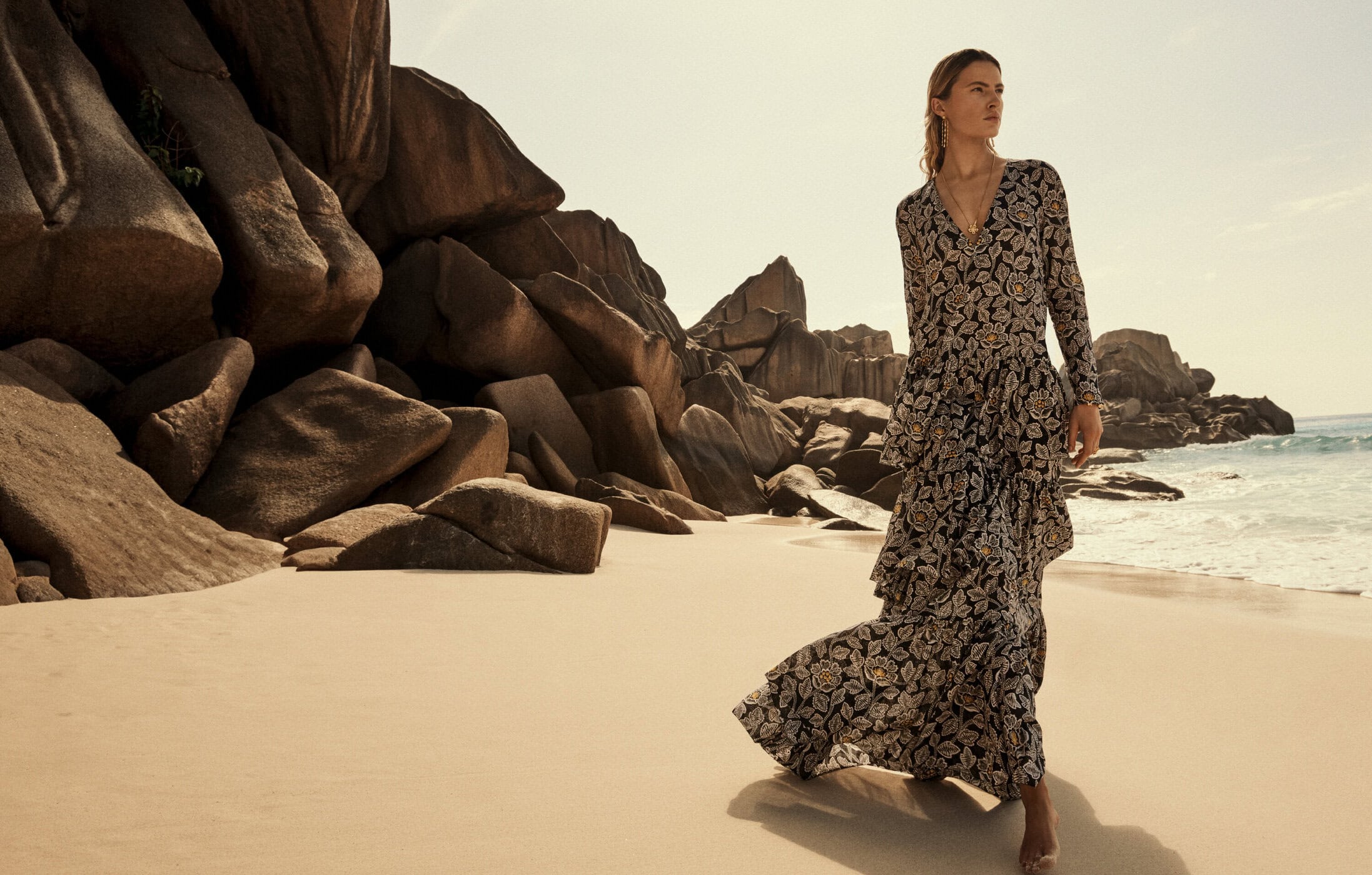 A blond woman in a floral dress walks on a sandy beach with granite rock formations under a bright, sunny sky and warm, soft light.