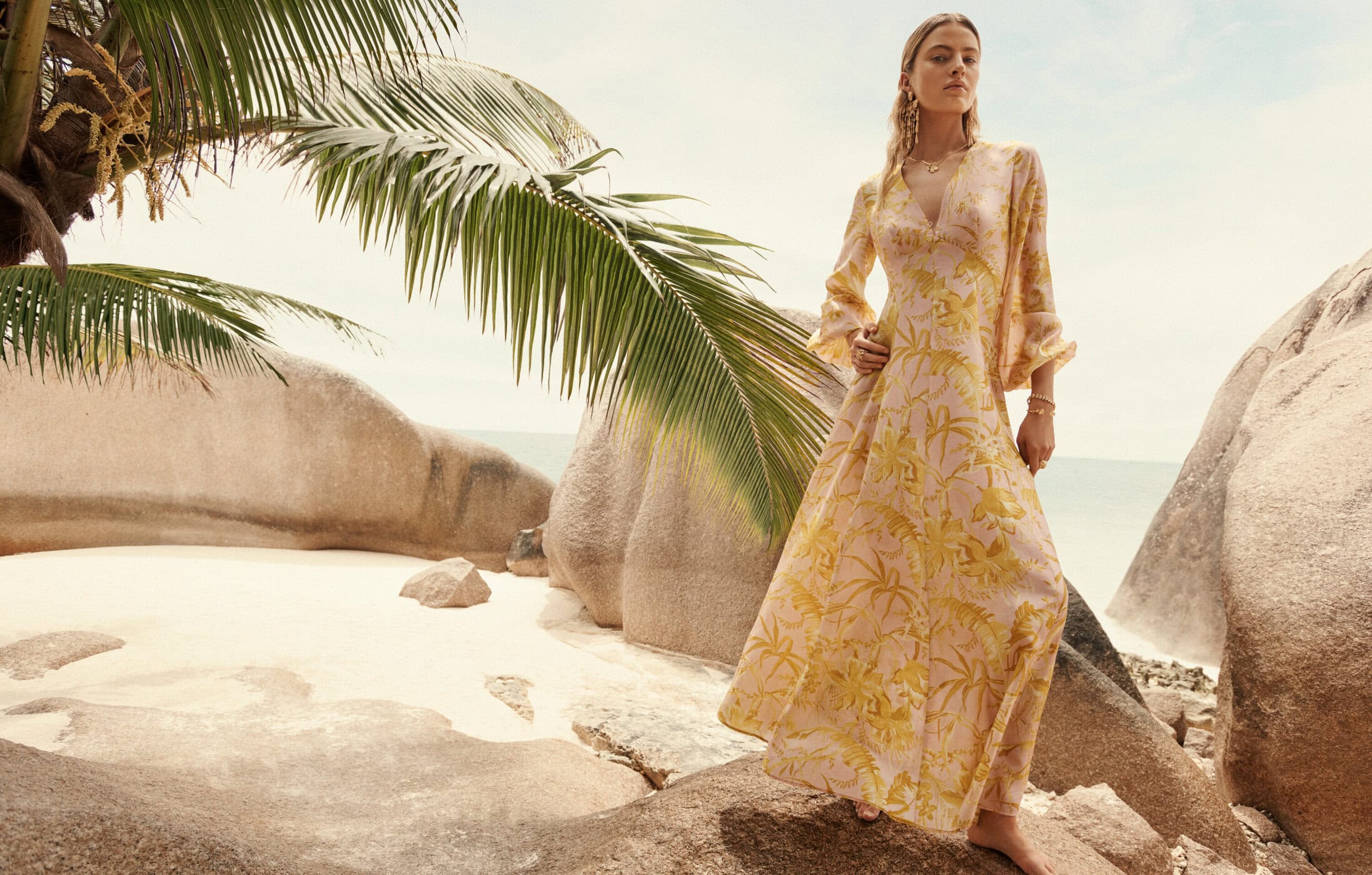 Young woman in flowing floral dress on a tropical beach with rock formations, palm trees, and blue sky. Fashion shot, outdoor setting.