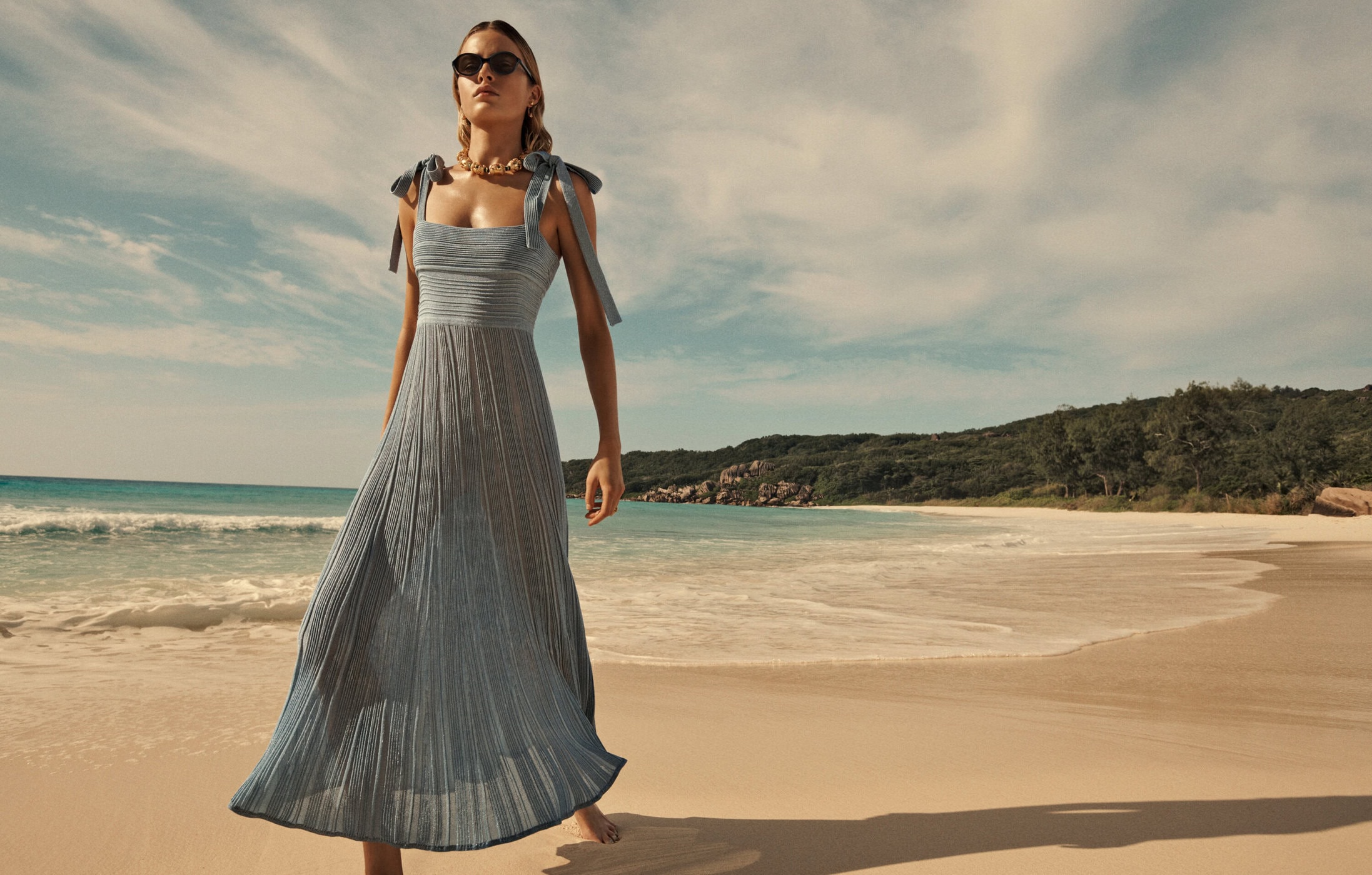A woman wearing a blue dress stands on a sandy beach with the ocean and a green treeline in the background on a sunny day.