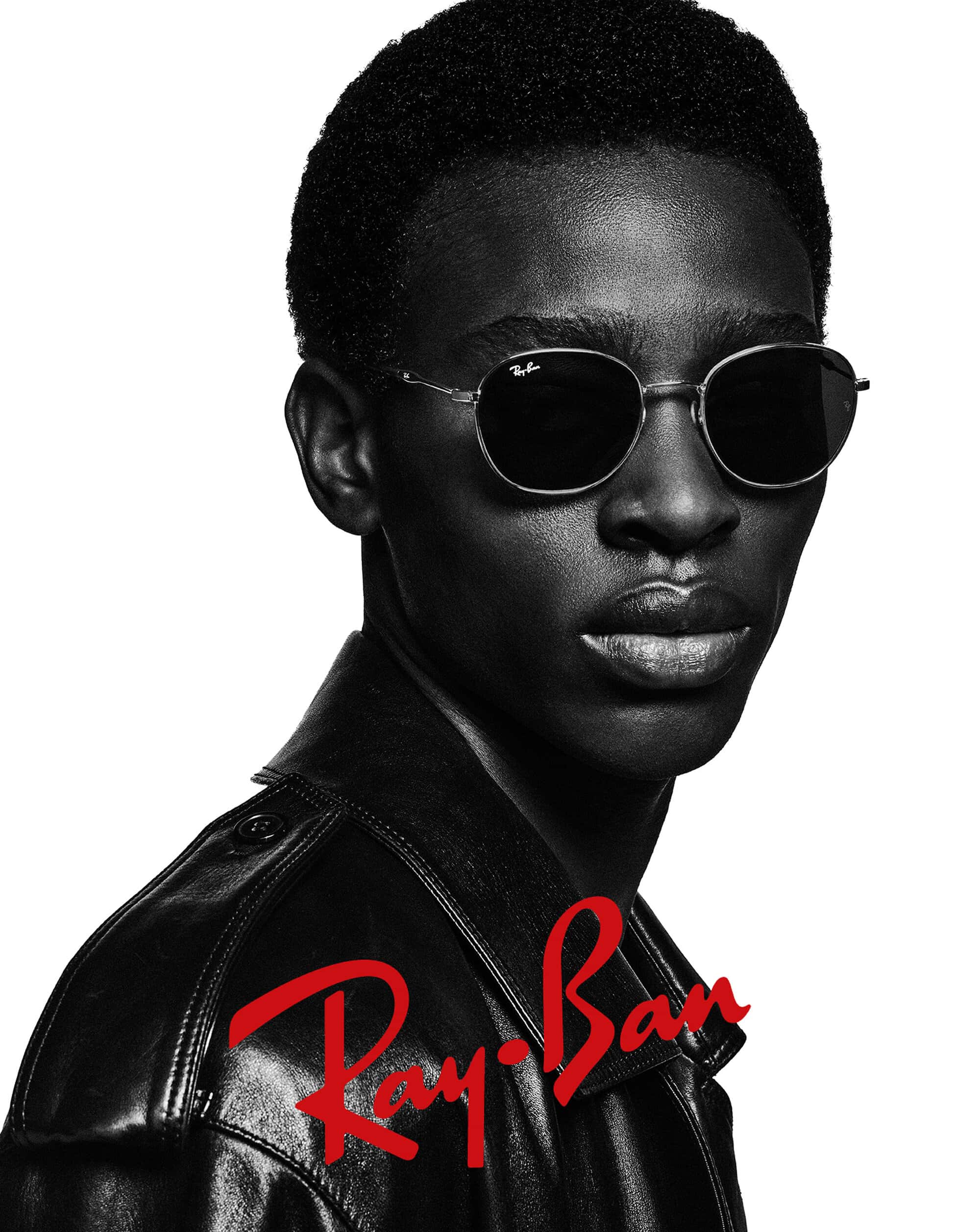 A close-up of a Black male model wearing Ray-Ban sunglasses and a black leather jacket against a white backdrop, showcasing fashion.