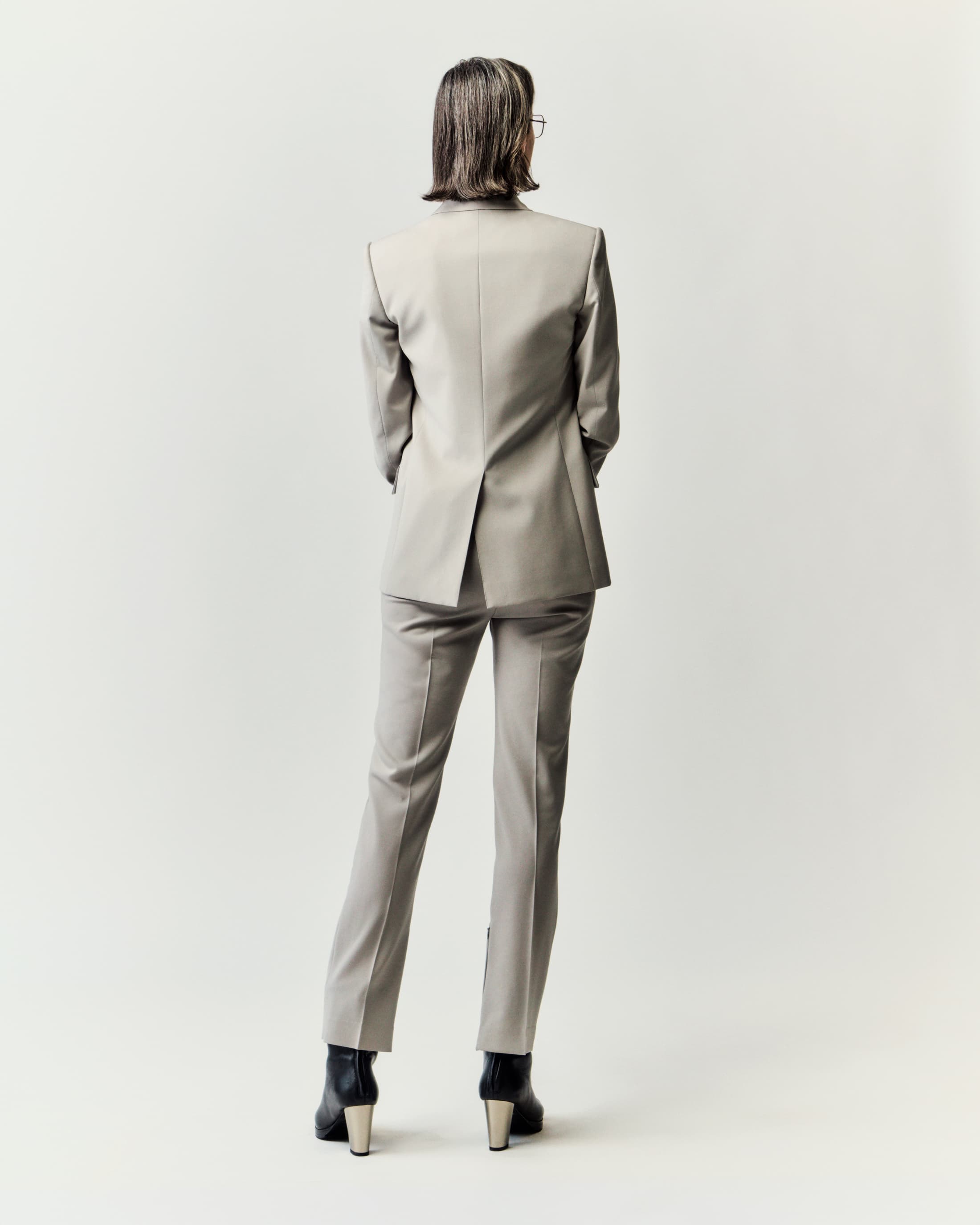 Rear view of an adult woman in a light grey suit with black boots, standing against a white backdrop, showcasing formal business attire.